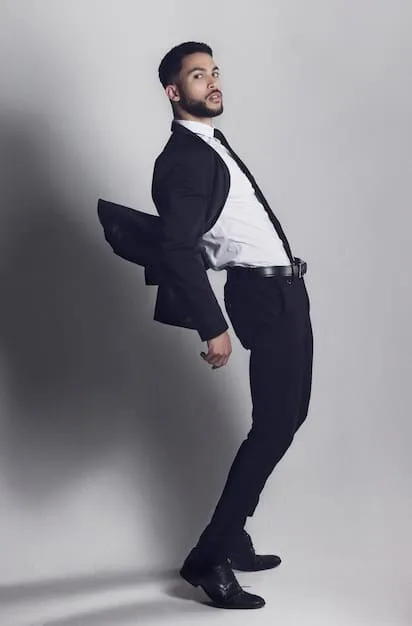 A full-length shot of a male celebrity wearing a suit with a unique, deconstructed design. The suit features visible stitching, raw edges, and an asymmetrical cut. He is accessorized with modern jewelry and minimalist shoes.